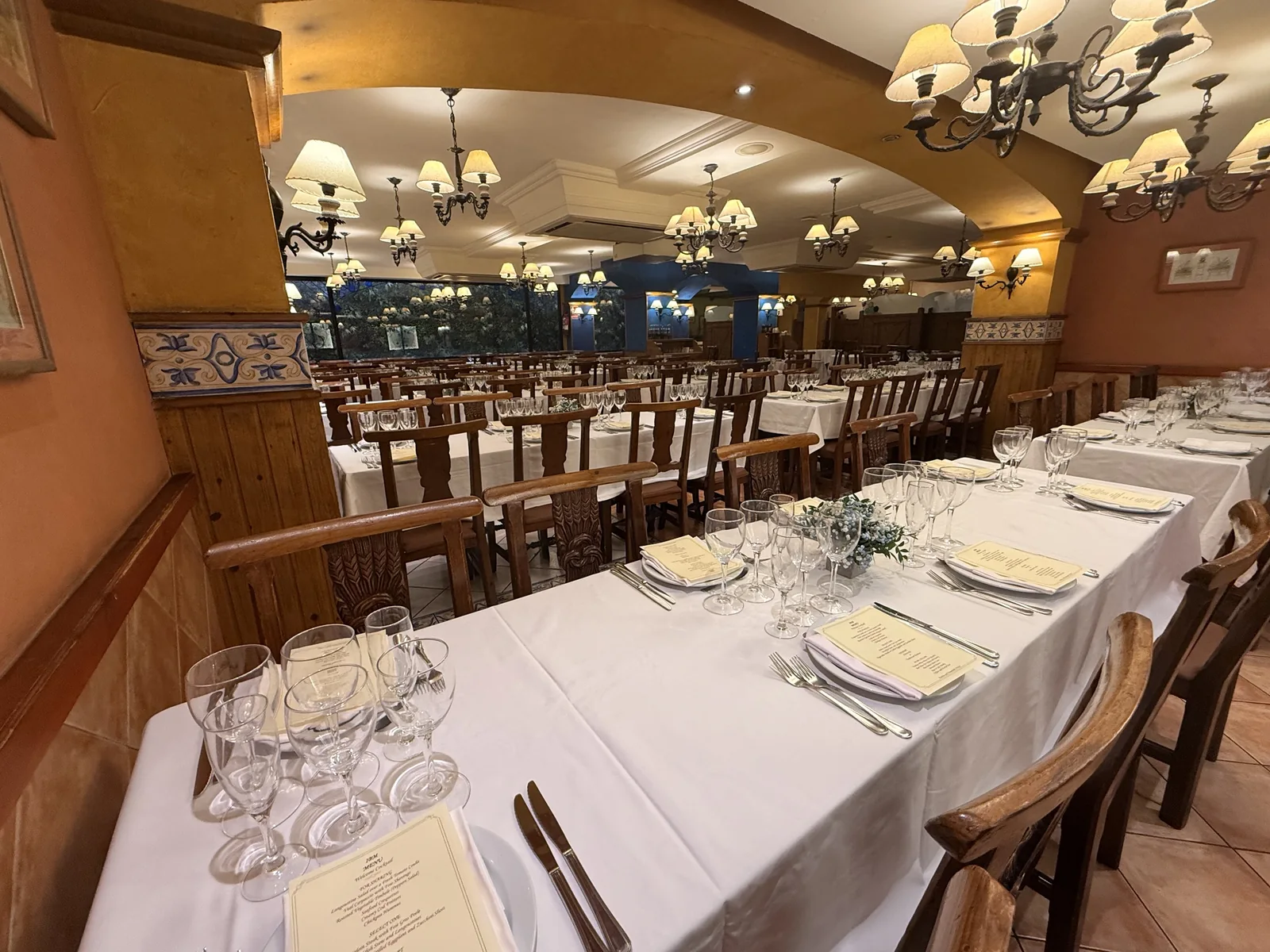 Mesa larga con mantel blanco, copas y sillas de madera bajo arcos y lámparas.