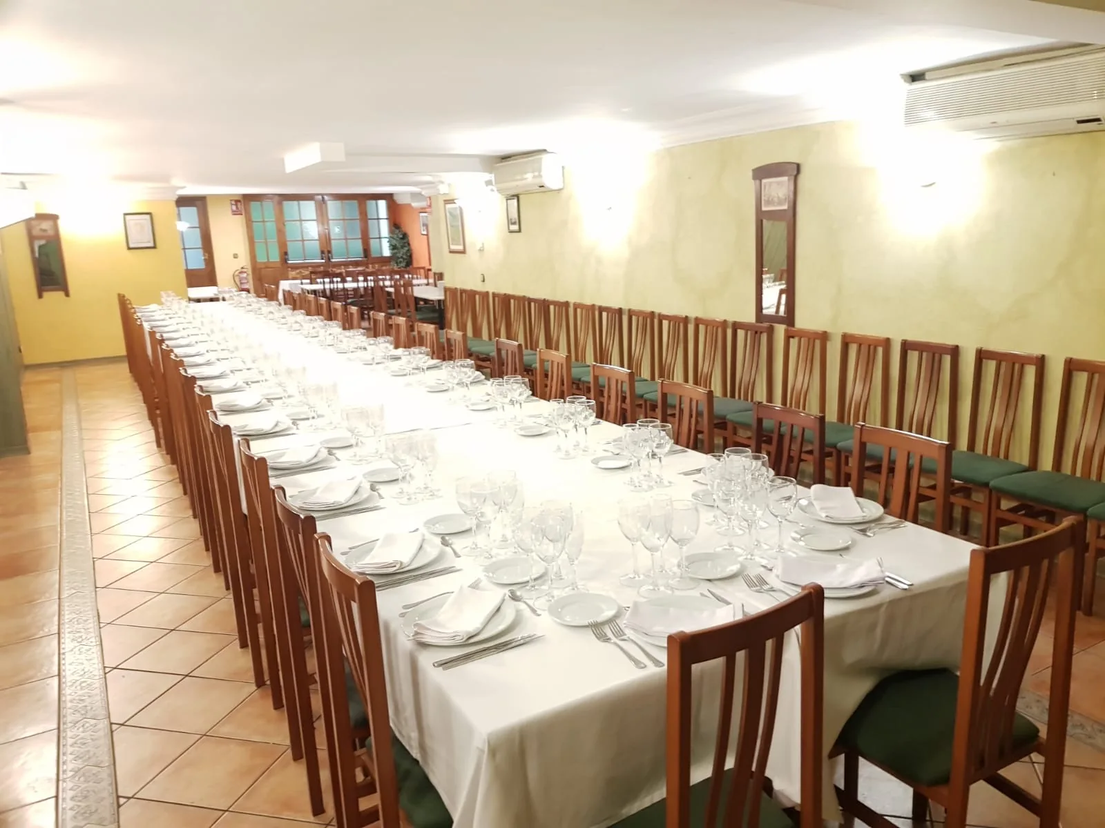 Salón de paredes claras con una mesa larga preparada con manteles blancos y sillas de madera.